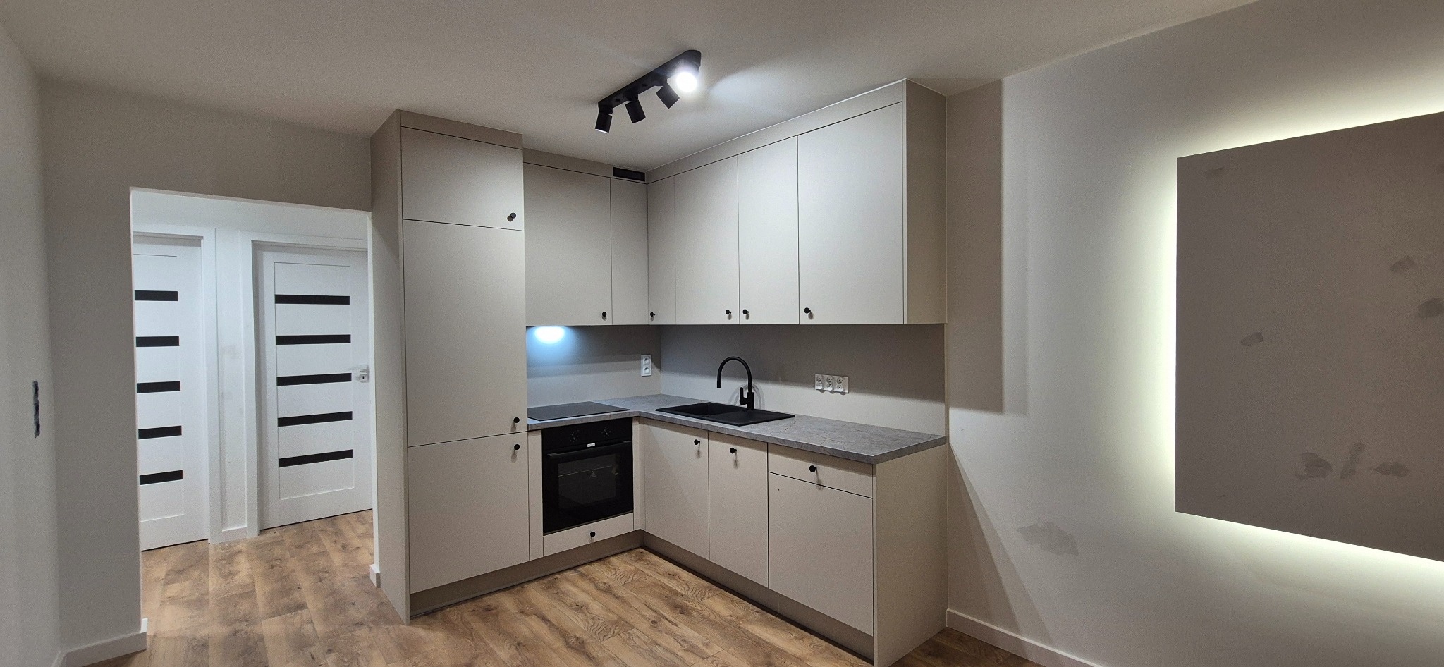 Jasna kuchnia na wymiar z czarnymi akcentami: minimalistyczne szafki, czarny zlew i bateria, oświetlenie punktowe. Widok na drewnopodobną podłogę i drzwi z czarnymi paskami.