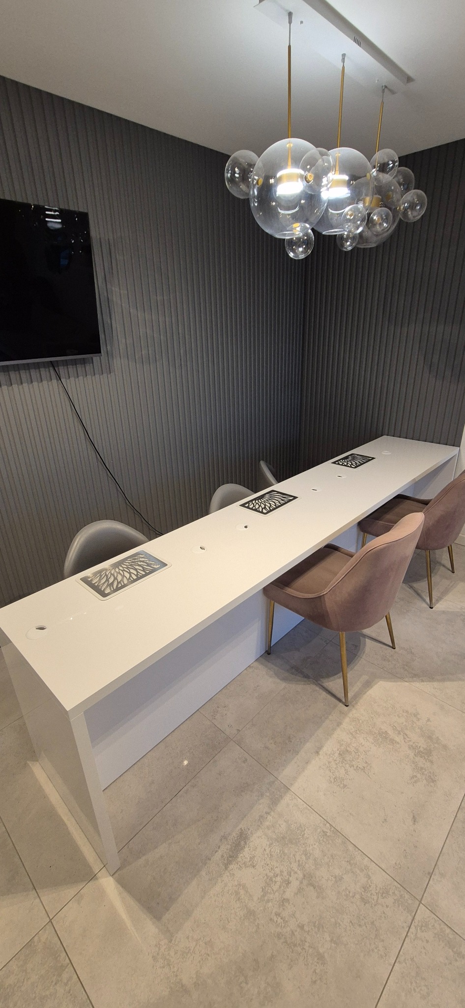 Długi, biały stół do manicure z wbudowanymi pochłaniaczami pyłu, oświetlony designerską lampą. W tle szara ściana z pionowymi żłobieniami i telewizor.