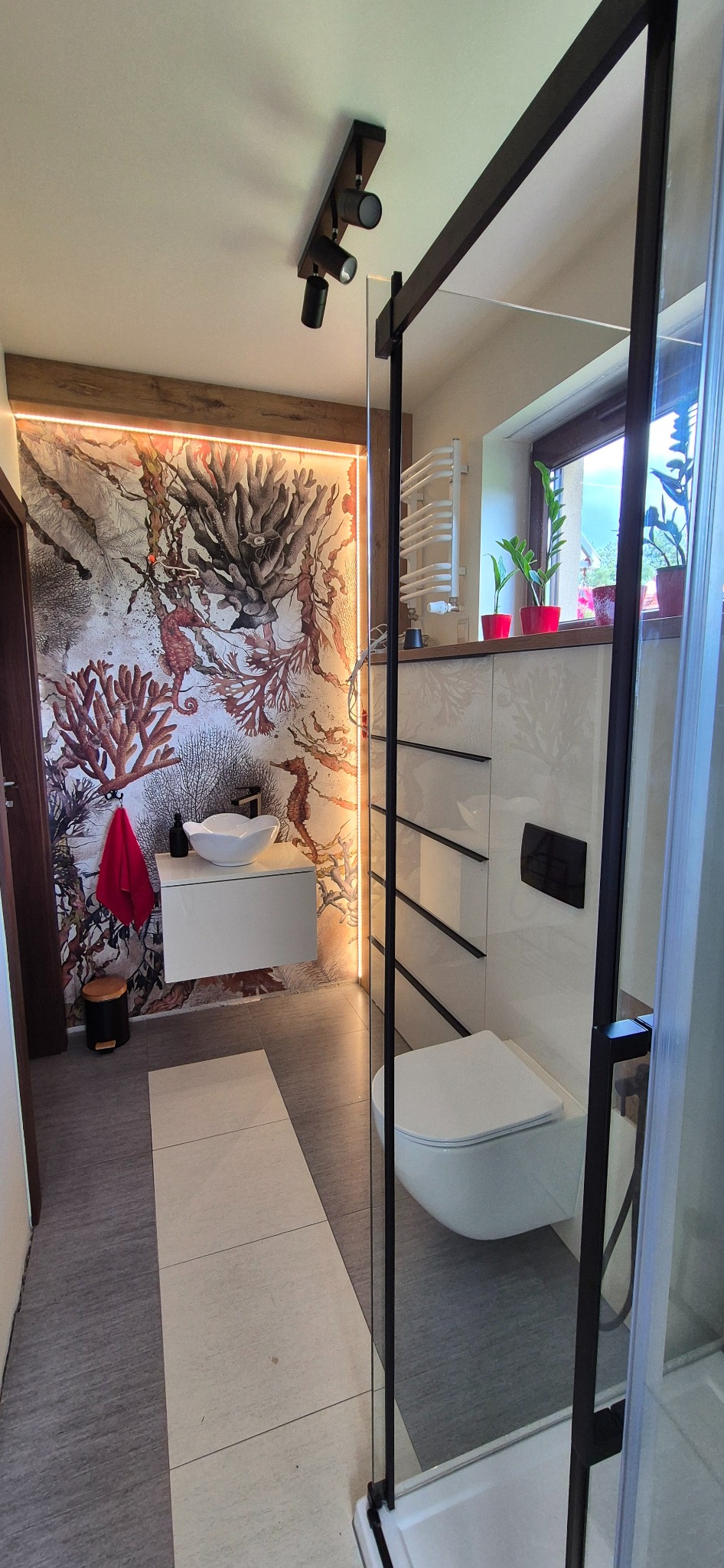 Nowoczesna łazienka z kabiną typu walk-in, umywalką na szafce i tapetą z motywem morskim. Oświetlenie punktowe i rośliny w doniczkach dodają uroku.