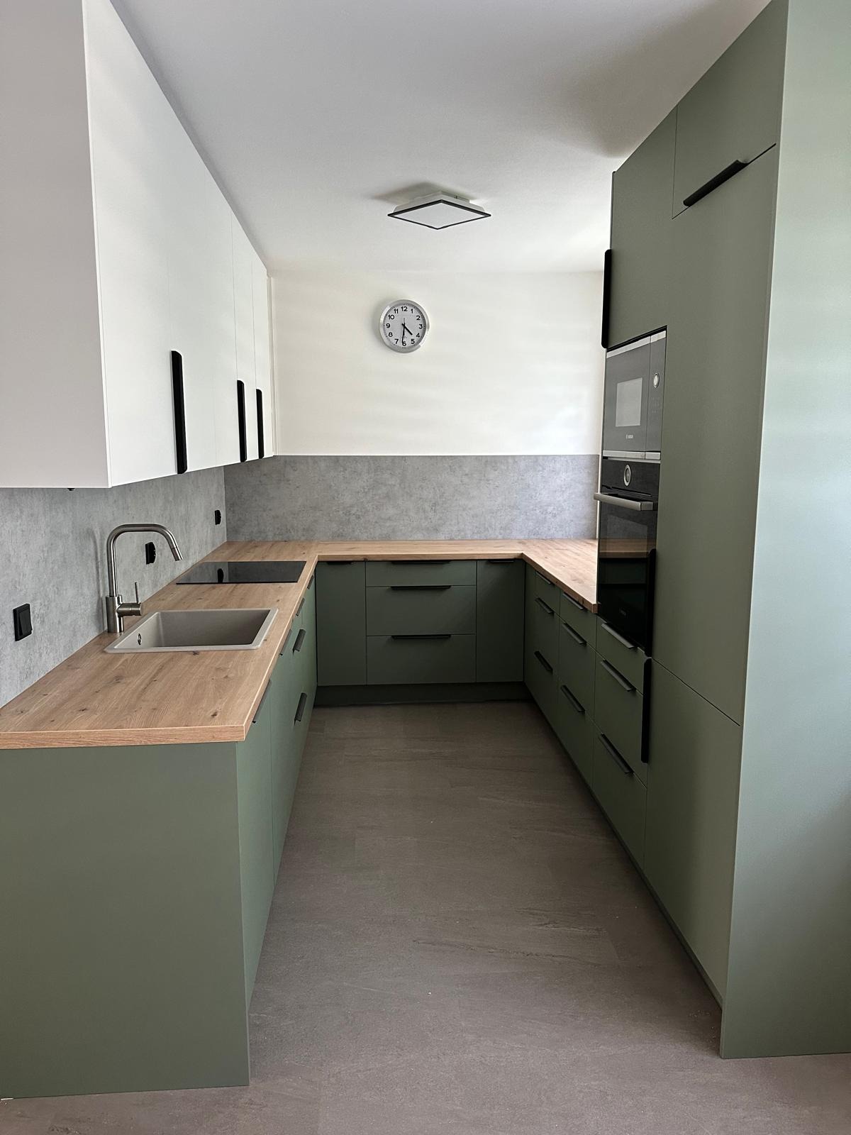 Nowoczesna kuchnia w kształcie litery U z oliwkowymi szafkami, drewnianym blatem i białymi szafkami wiszącymi. Minimalistyczny design z czarnymi uchwytami i zegarem na ścianie.