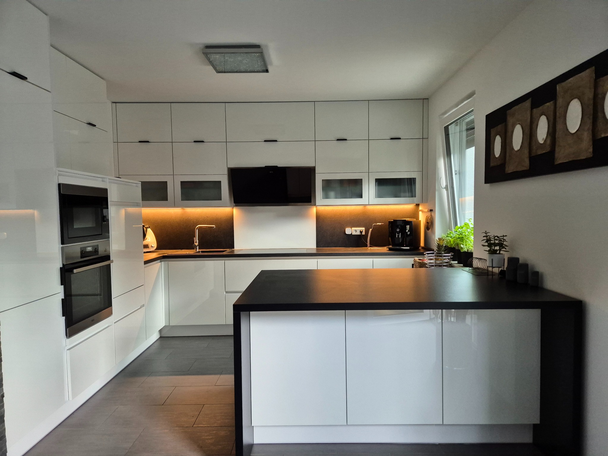 Biała, nowoczesna kuchnia z wyspą i czarnym blatem, oświetleniem LED pod szafkami, minimalistycznymi uchwytami i dekoracją ścienną w kształcie owali.