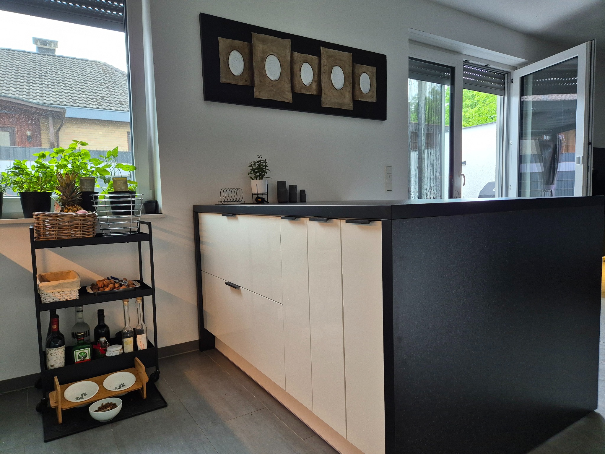 Nowoczesna wyspa kuchenna z białymi frontami i czarnym blatem, uzupełniona dekoracjami i roślinami w tle, tworzy przytulną atmosferę wnętrza.