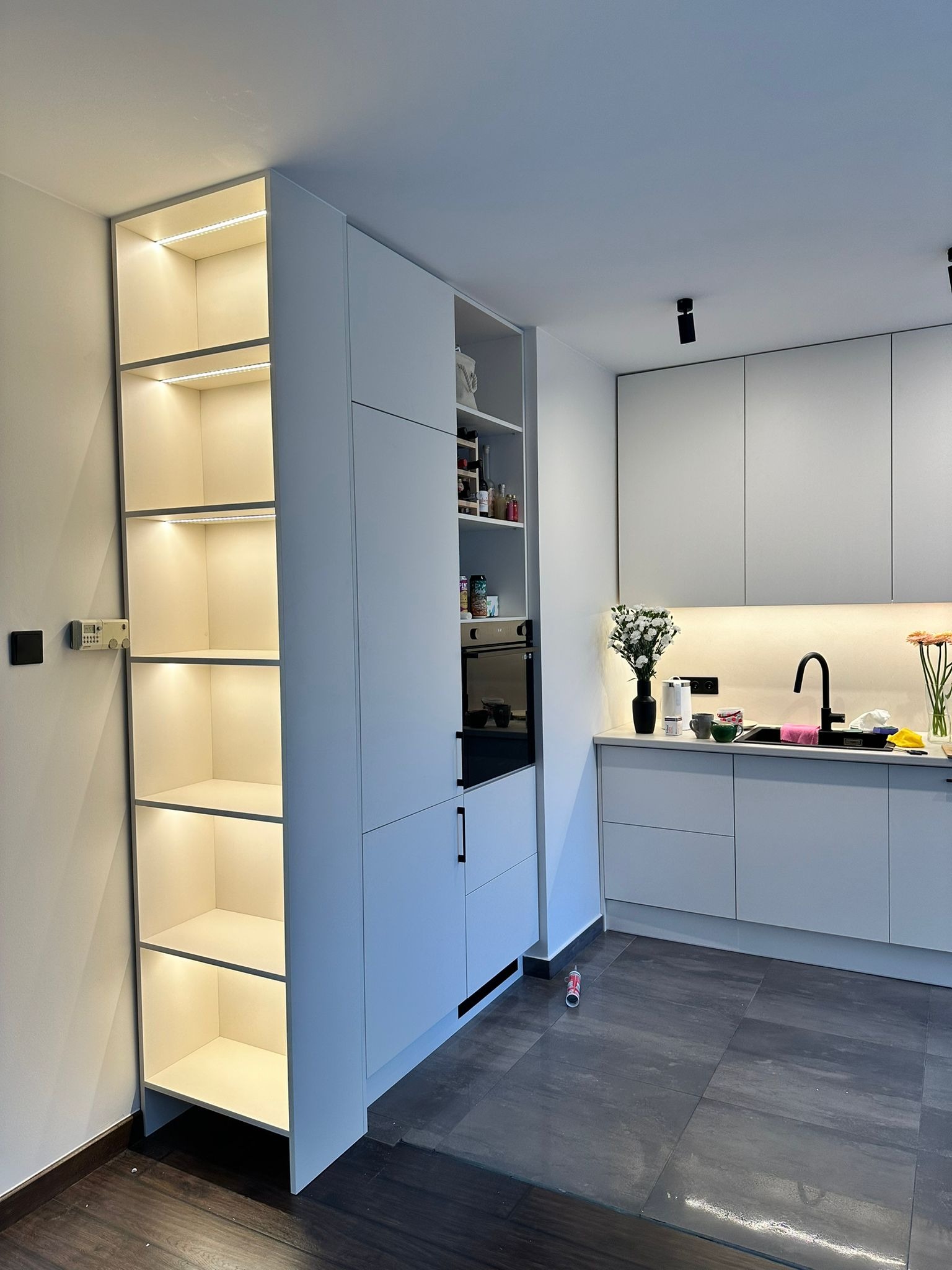 Nowoczesna, jasna kuchnia z zabudową na wymiar: szafki z oświetleniem LED, piekarnik w zabudowie i minimalistyczny design. Ciemna podłoga z płytek.