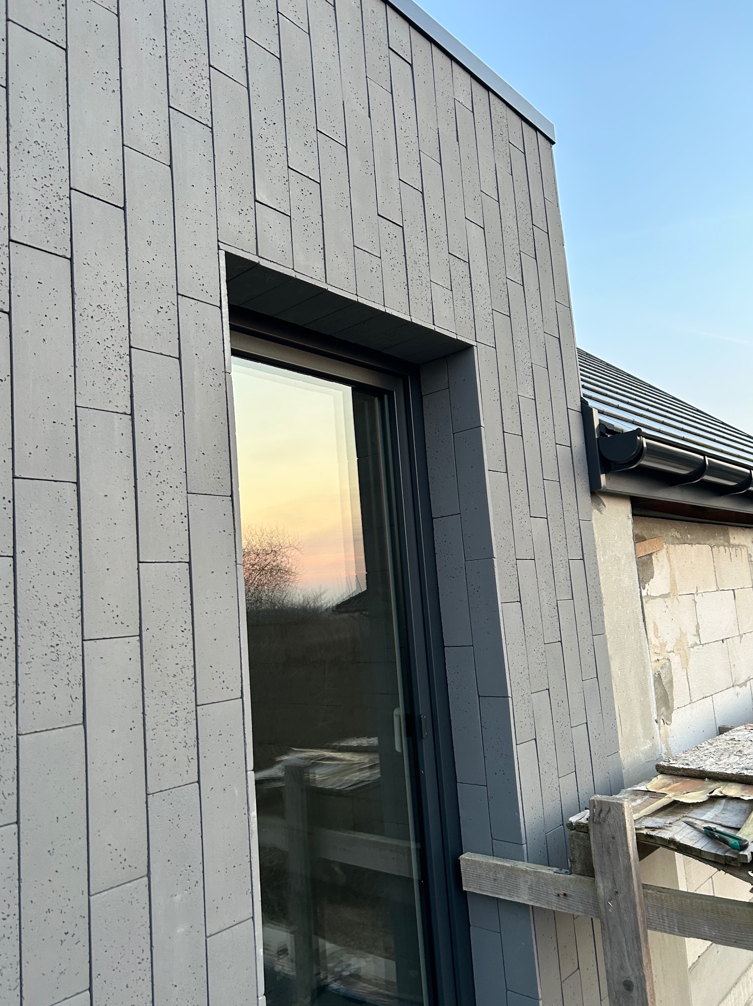 Fragment elewacji budynku z szarej, podłużnej płytki imitującej kamień, widoczne okno z odbijającym się zachodem słońca oraz fragment orynnowania dachu i rusztowania.