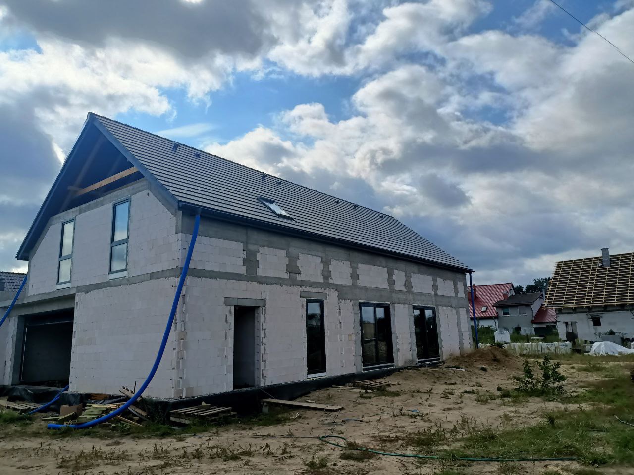Dom w trakcie budowy z szarą dachówką, widoczne okna i drzwi, nieotynkowane ściany z betonu komórkowego, niebieskie rury na zewnątrz budynku.