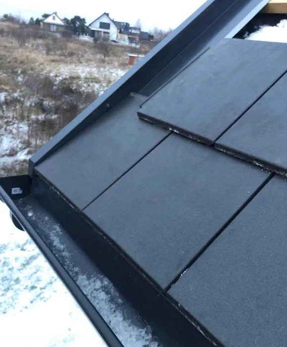 Fragment dachu pokrytego ciemnoszarą dachówką ceramiczną, z widoczną rynną wypełnioną resztkami śniegu i ośnieżonym tłem z zabudowaniami.