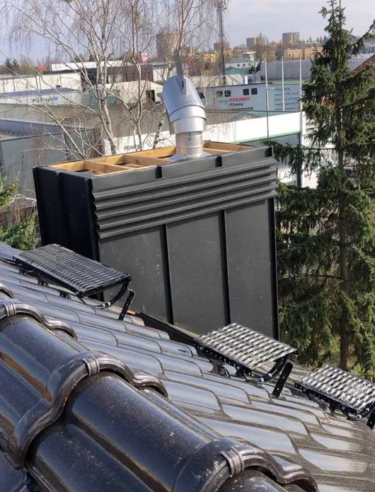 Błyszcząca dachówka ceramiczna z zamontowanymi stopniami kominiarskimi i nowym kominem z metalową nasadą, na tle zabudowań miejskich.