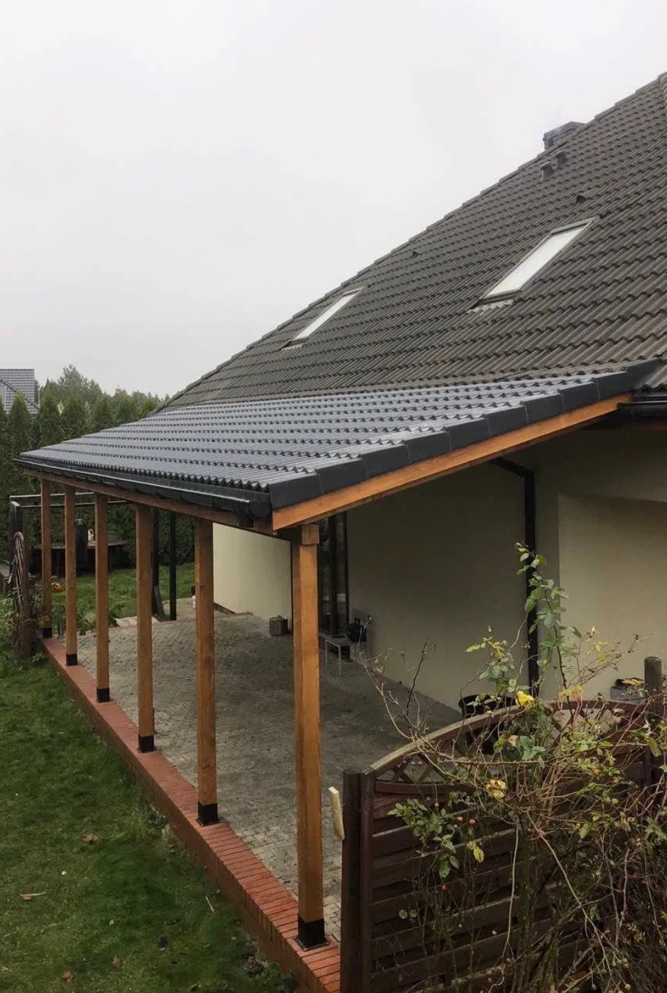 Zadaszenie tarasu z drewnianymi słupami i nowym pokryciem z blachy trapezowej, widok na dach domu z dwoma oknami dachowymi w pochmurny dzień.