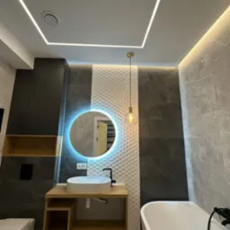 Nowoczesna łazienka z okrągłym lustrem podświetlanym LED, wiszącą szafką z umywalką nablatową, białą wanną i dekoracyjną ścianą z trójwymiarowych płytek.