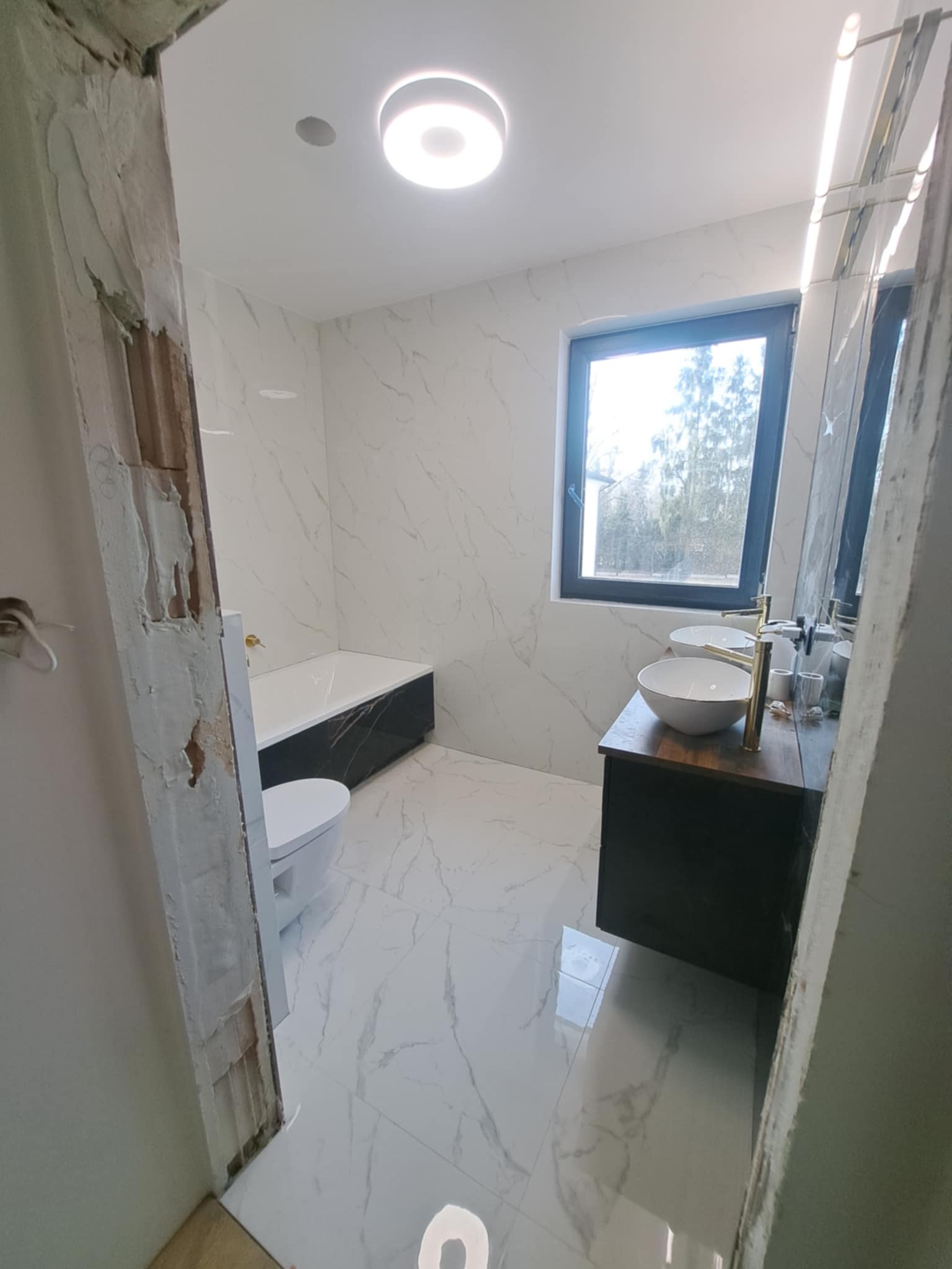 Wnętrze łazienki w trakcie remontu: widoczna wanna z czarną obudową, toaleta, umywalka z dwoma złotymi kranami, okno z widokiem na drzewa oraz fragment ościeżnicy w stanie surowym.