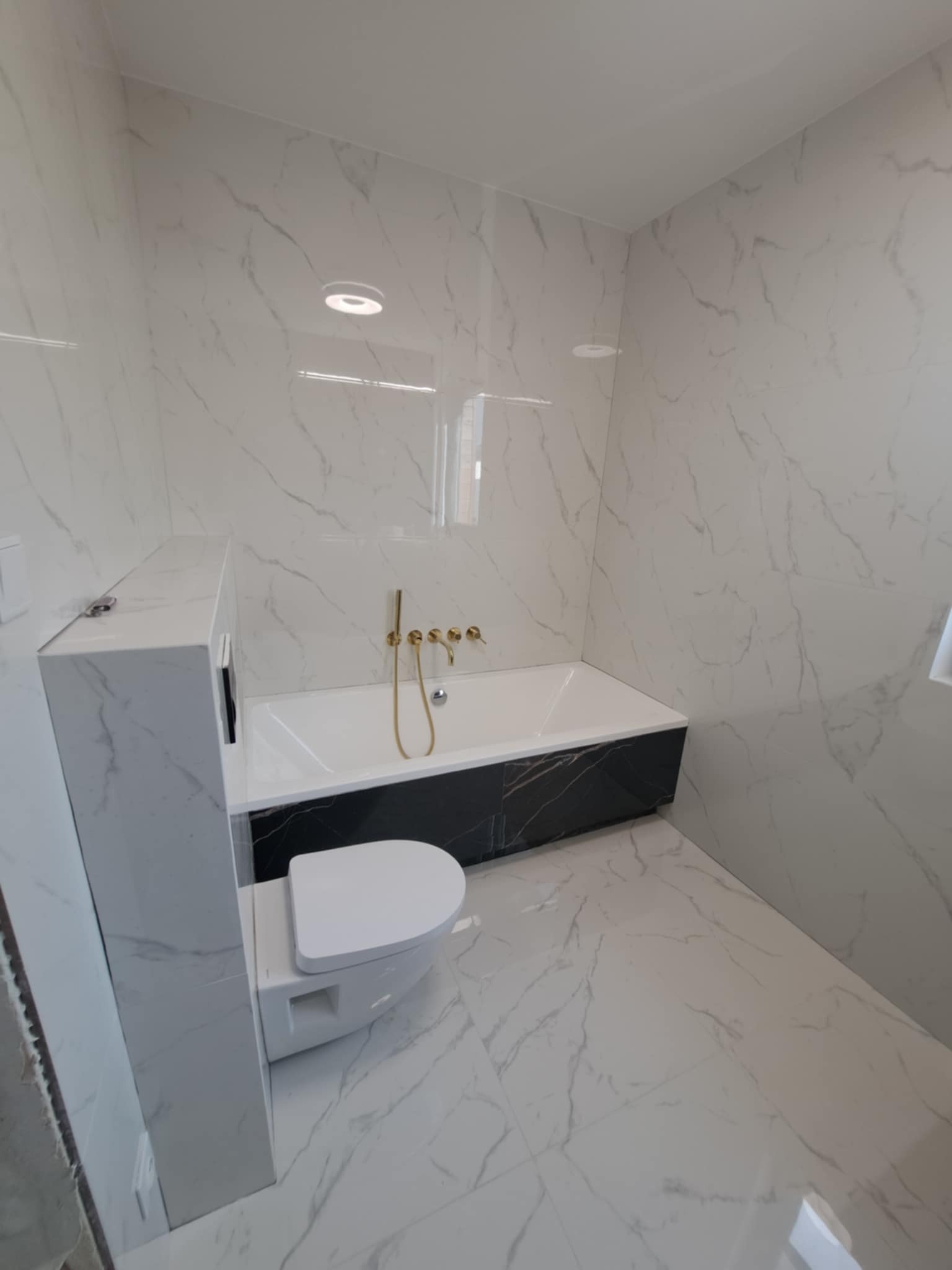 Nowoczesna łazienka z białymi marmurkowymi płytkami, czarną obudową wanny i złotą armaturą, toaleta w zabudowie.