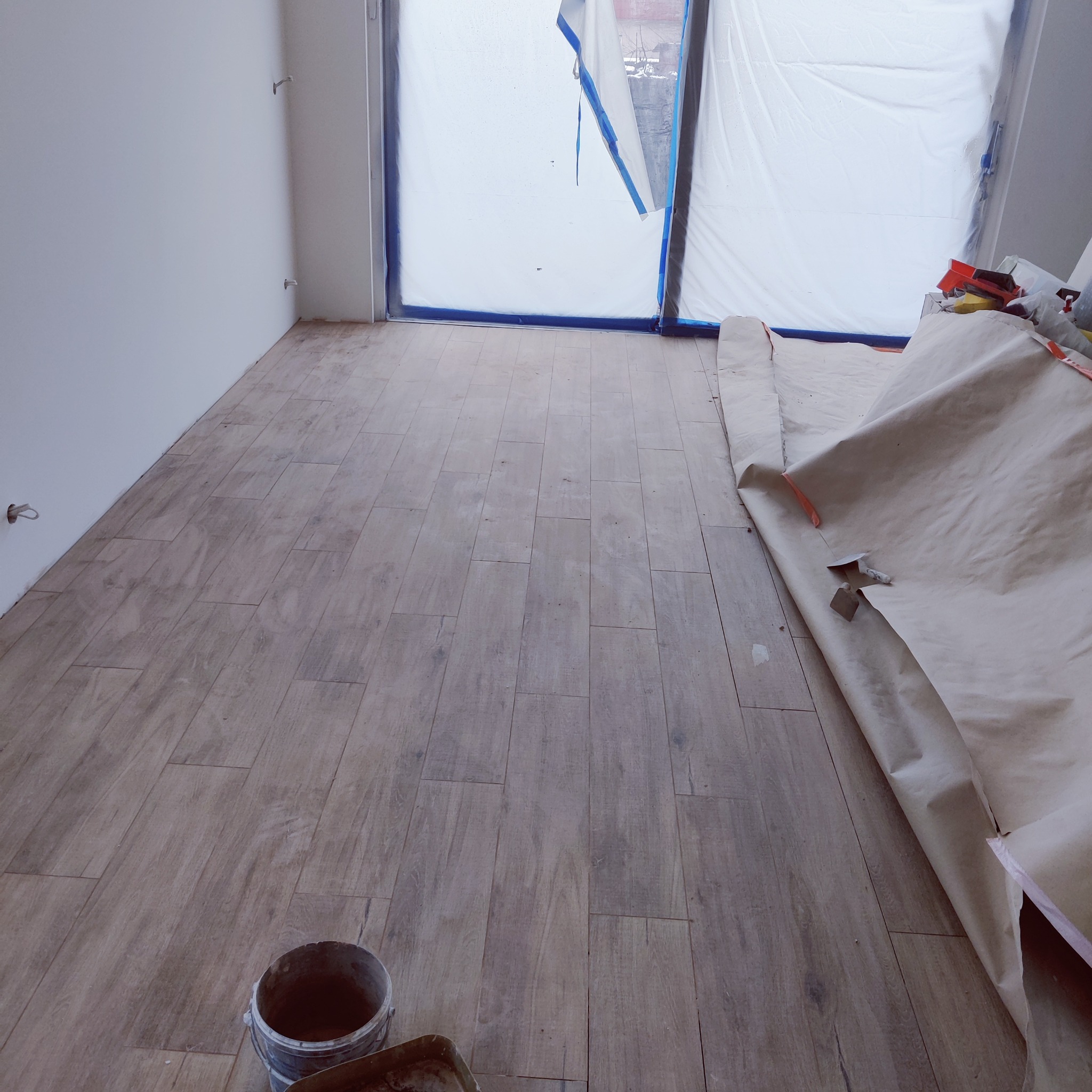 Ułożona podłoga z płytek imitujących drewno w trakcie remontu, zabezpieczone okno folią malarską, widoczne wiadro i kuweta glazurnicza.