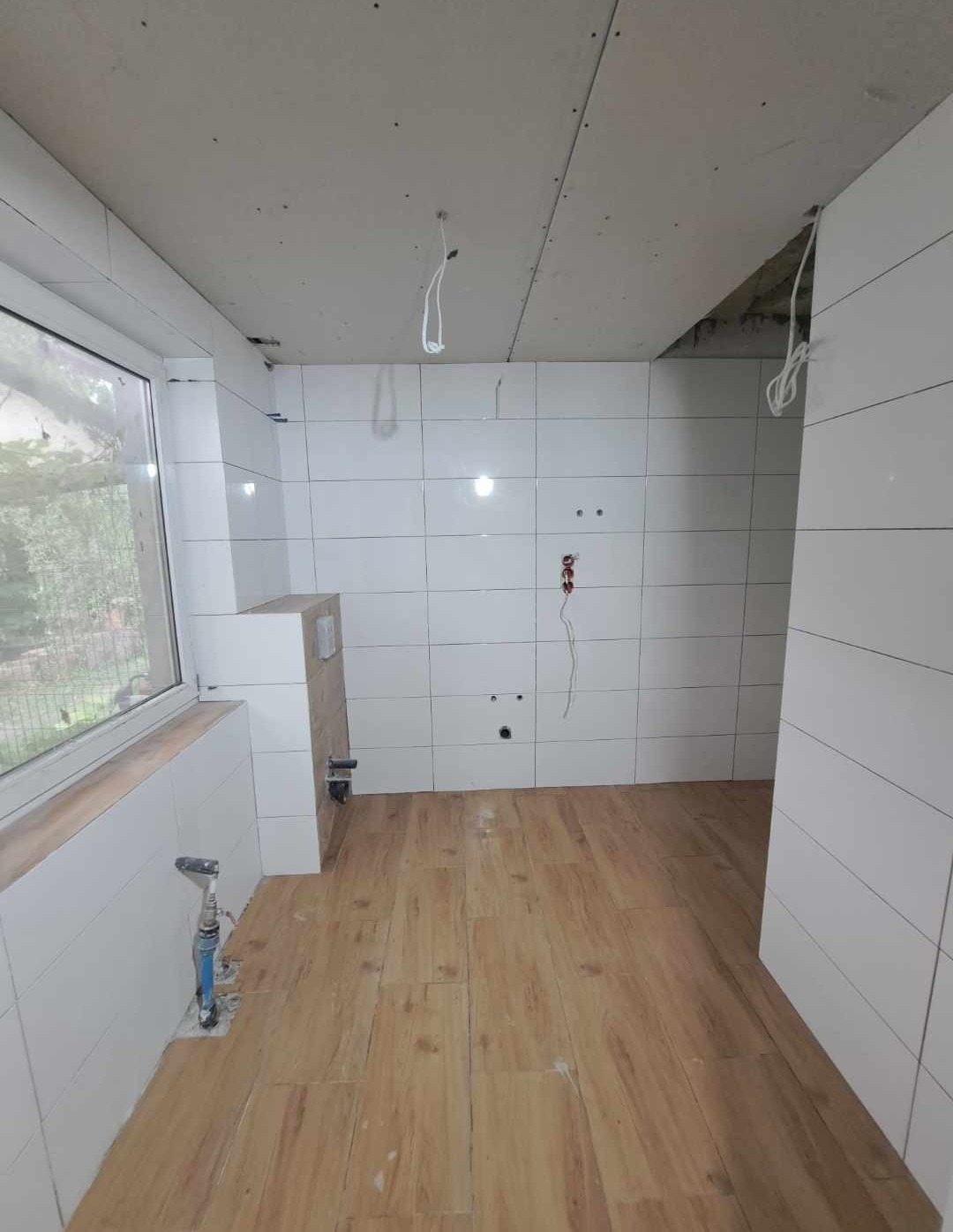 Łazienka w trakcie remontu: białe płytki na ścianach, drewnopodobne panele na podłodze, surowy sufit z płyt gipsowo-kartonowych, wystające kable elektryczne i instalacja wodna przed zabudową.