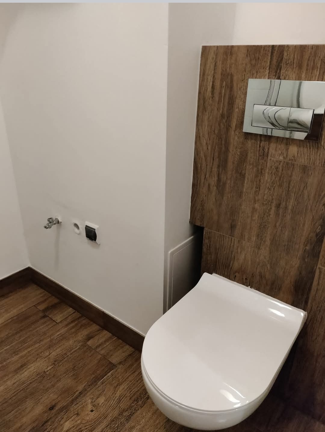 Wisząca toaleta z chromowanym przyciskiem spłukującym na tle drewnopodobnych płytek, widoczne przyłącza wodne i elektryczne na ścianie, podłoga wykończona panelami imitującymi drewno.