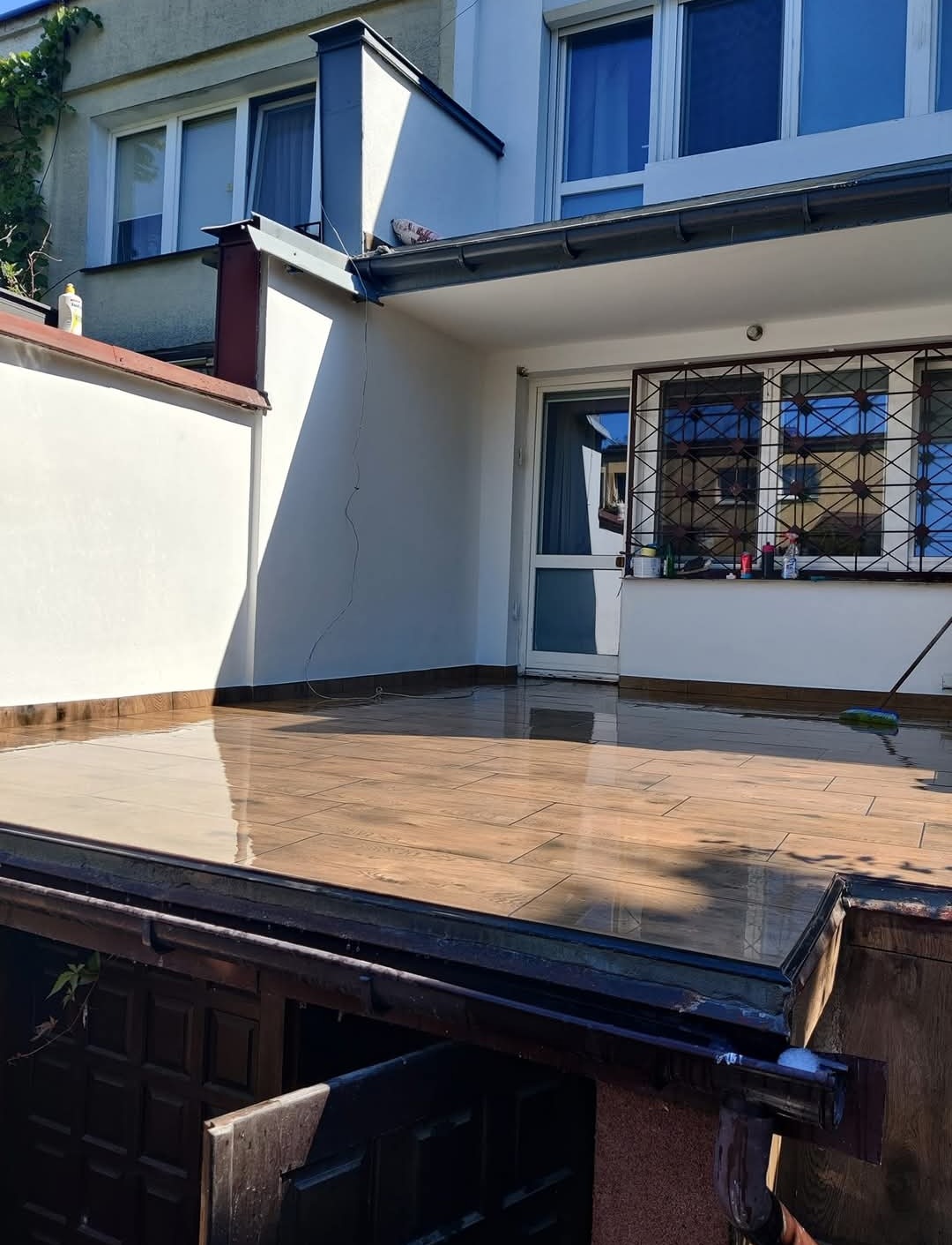 Błyszczący taras wyłożony brązowymi płytkami imitującymi drewno, po umyciu, z widocznym odbiciem nieba i fragmentem elewacji budynku z białymi ścianami i oknami z ozdobną kratą.