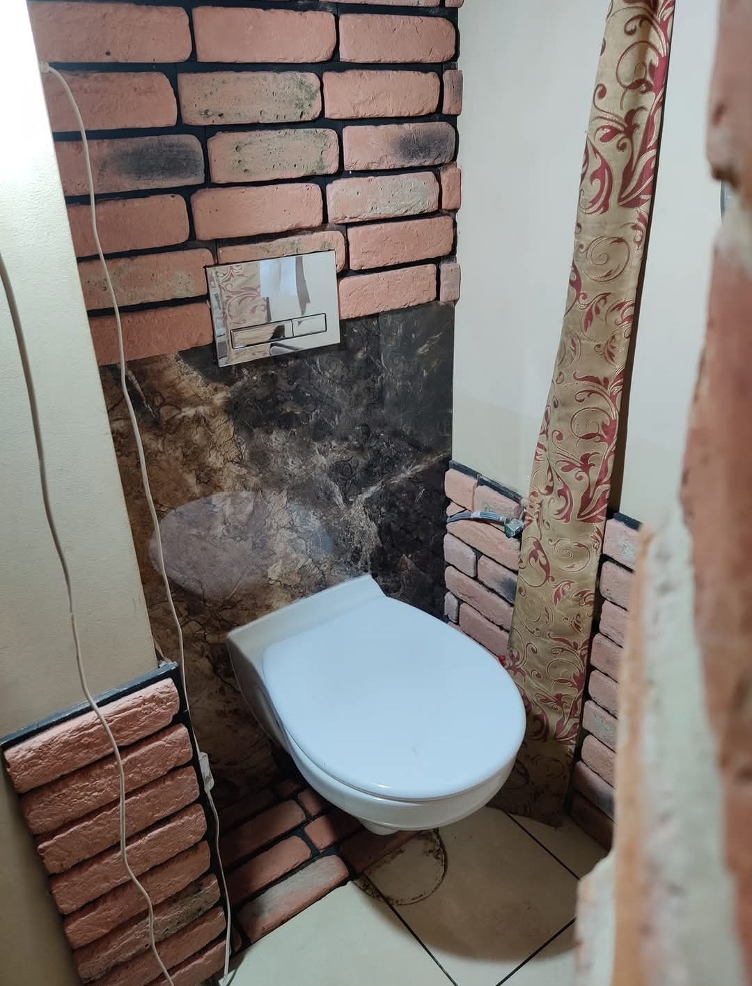 Toaleta podwieszana w łazience z częściowo wykończonymi ścianami imitującymi cegłę, widoczna instalacja spłuczki i fragment zasłony z motywem roślinnym.