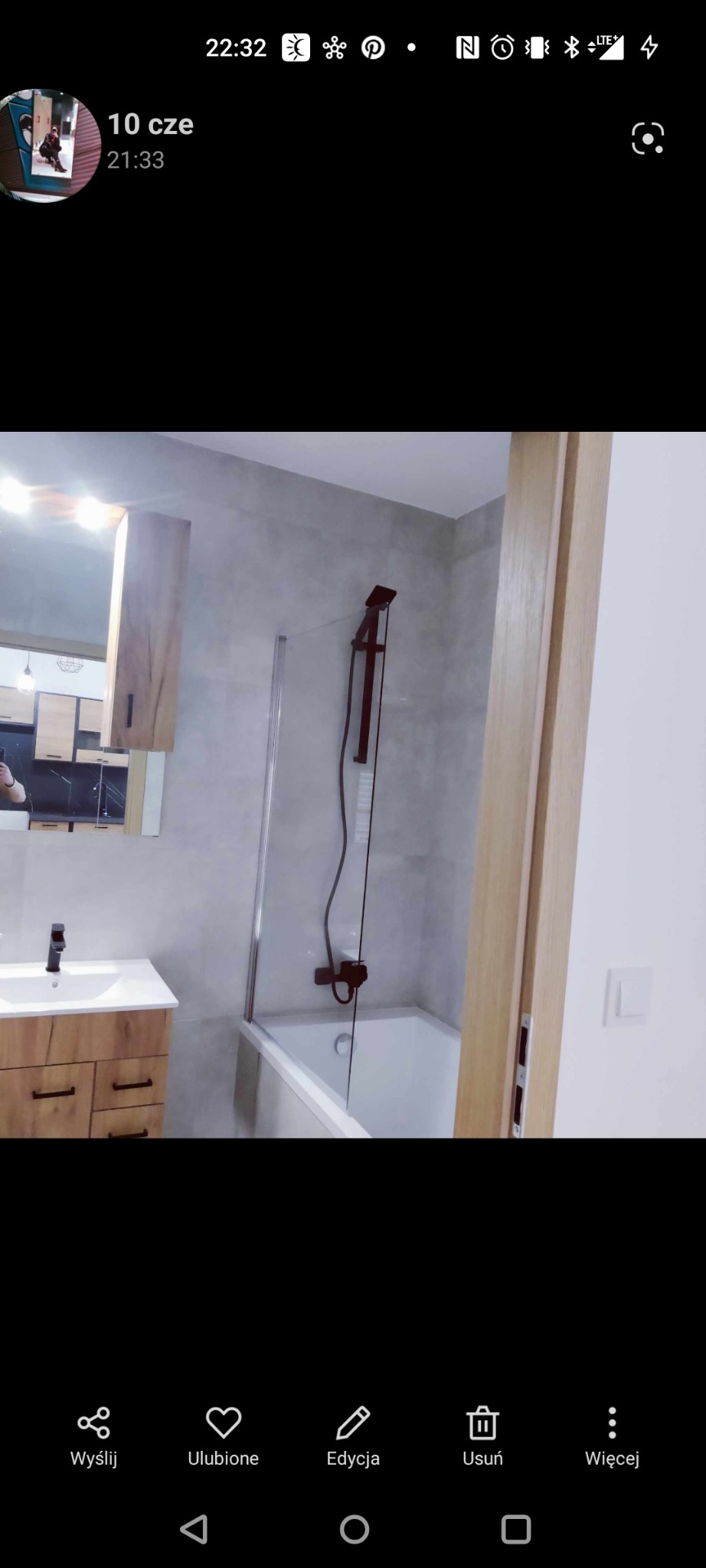 Nowoczesna łazienka z wanną i szklanym parawanem, umywalka z szafką w kolorze drewna, ciemna armatura, szare płytki na ścianach, widok na fragment kuchni w tle.