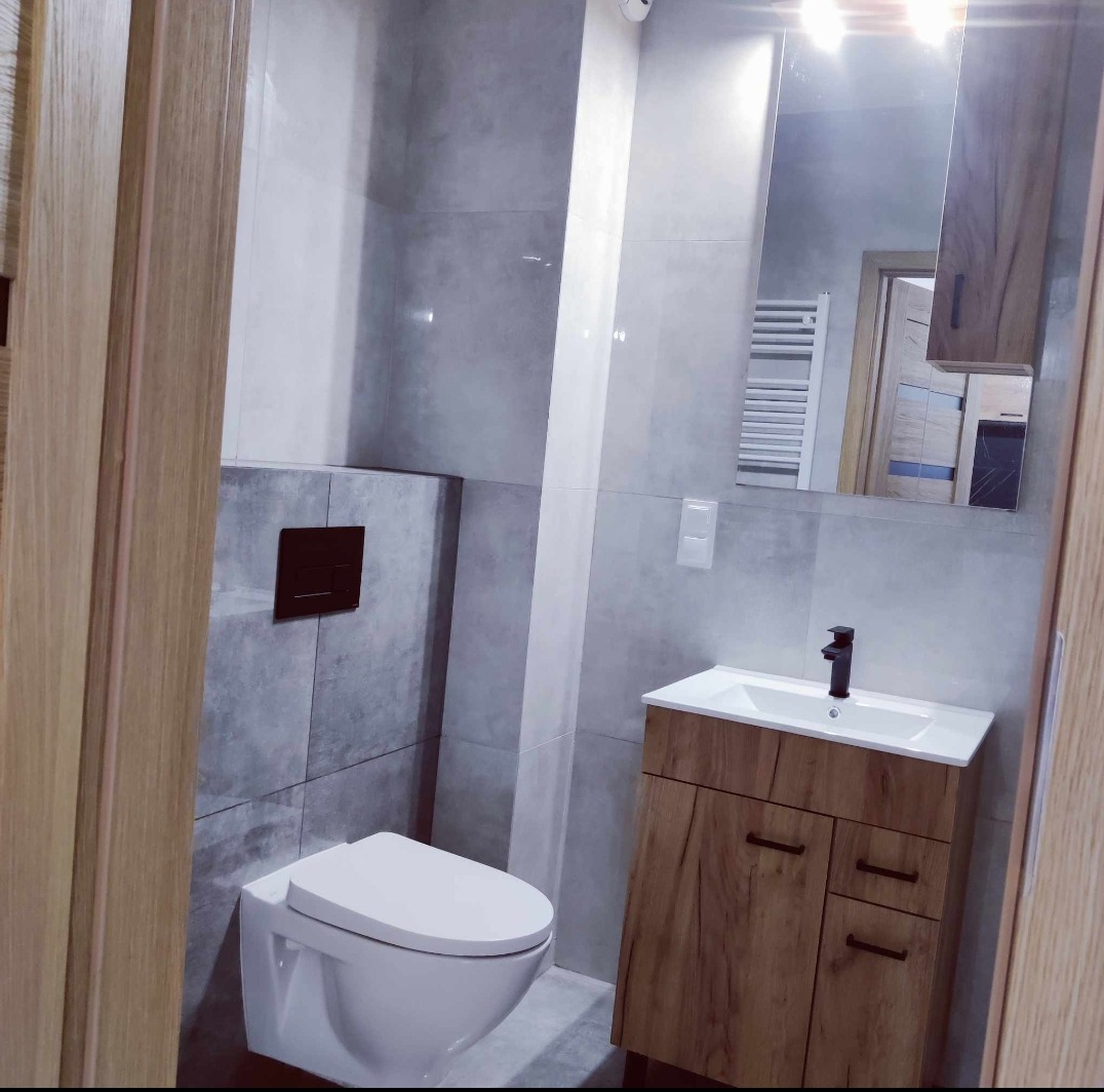 Wykończona łazienka w szarościach z drewnianą szafką pod umywalką i wiszącą toaletą z brązowym przyciskiem spłukiwania.