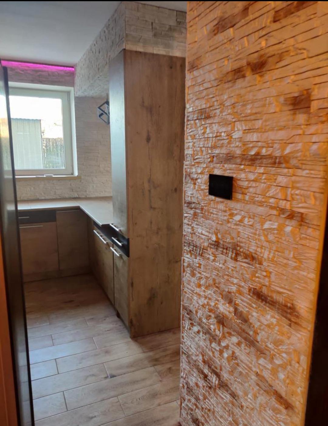 Wykończone wnętrze kuchni z drewnianymi meblami, podłogą z paneli oraz ścianą wyłożoną dekoracyjną cegłą w odcieniach brązu i beżu, z widocznym czarnym włącznikiem światła.