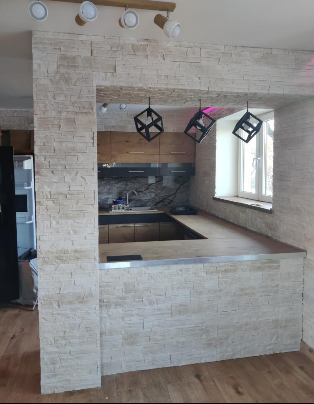 Wykończona kuchnia z barkiem obłożonym dekoracyjną, jasną cegłą, nowoczesne lampy wiszące i drewniane blaty.