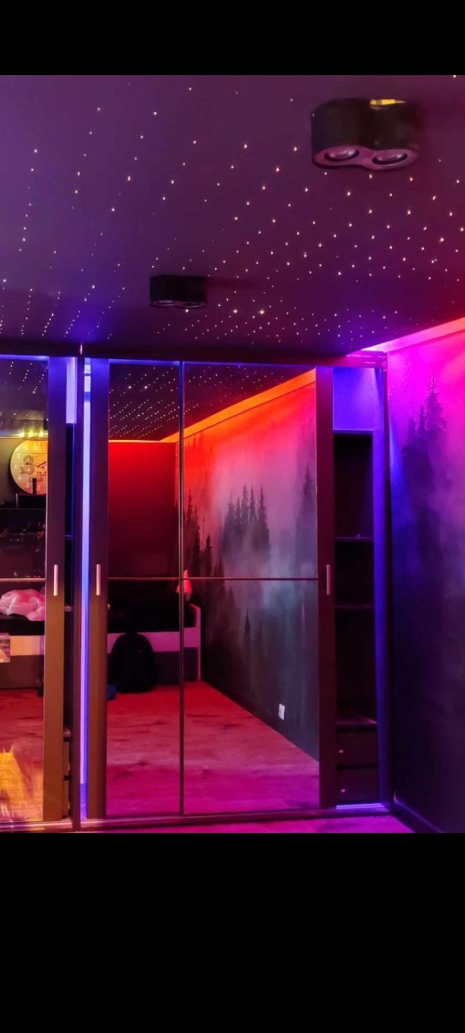 Pokój z szafą przesuwną z lustrami, podświetlonymi krawędziami, fototapetą z motywem lasu i sufitem imitującym gwieździste niebo, oświetlony kolorowymi lampami LED.