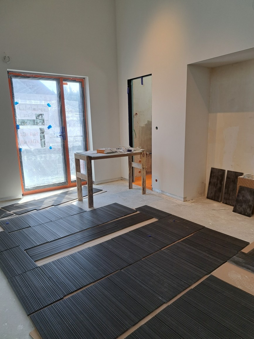 Ciemne, podłużne płytki ułożone częściowo na podłodze w pomieszczeniu w trakcie prac wykończeniowych; widoczna drabina malarska i okno zabezpieczone folią.