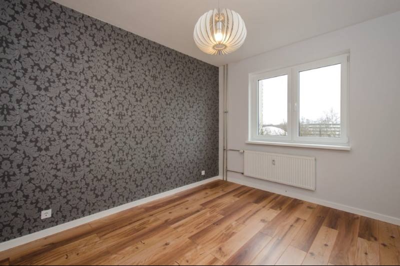 Pokój po remoncie: ściana z wzorzystą tapetą w odcieniach szarości, drewniana podłoga, białe okno i designerska lampa wisząca.