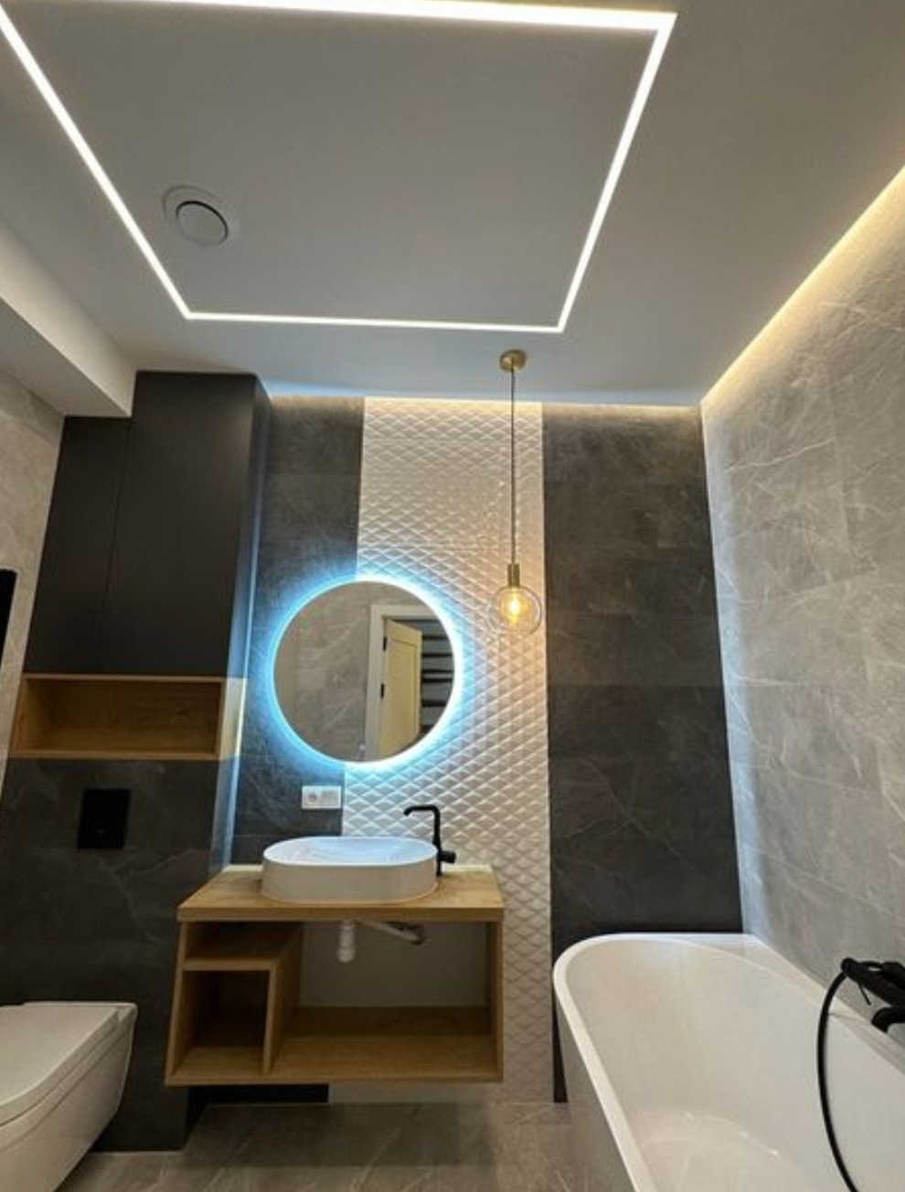 Nowoczesna łazienka z okrągłym lustrem podświetlanym LED, wiszącą szafką z umywalką nablatową, białą wanną i dekoracyjną ścianą z trójwymiarowych płytek.