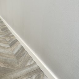 Sebastian Klimczak - Fragment pokoju po remoncie: jasnoszara ściana pomalowana farbą o matowym wykończeniu, biała listwa przypodłogowa i ułożone panele podłogowe w wzór jodełki francuskiej, imitujące drewno.