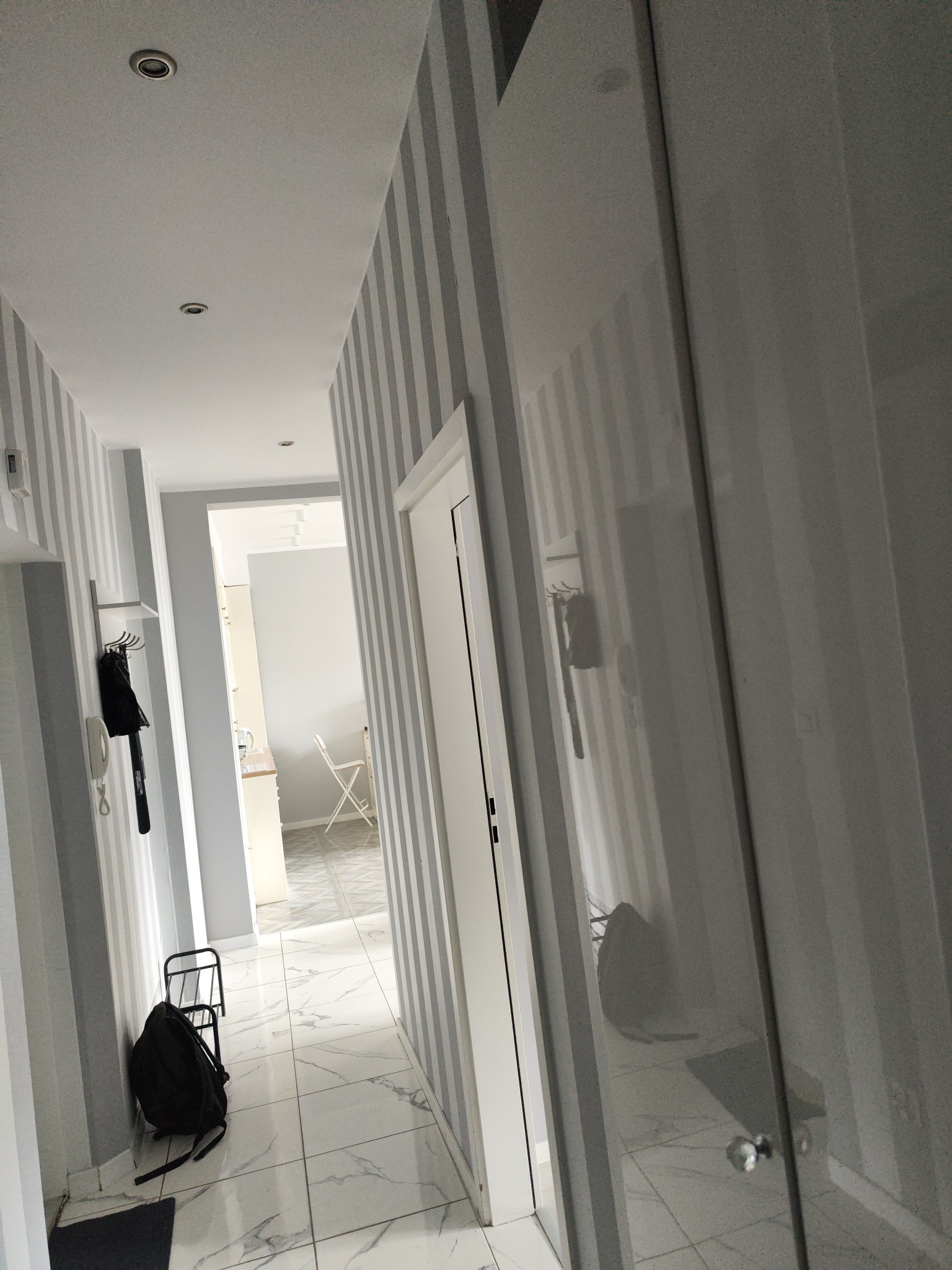 Korytarz z szaro-białymi pasami na ścianach i marmurową podłogą, widoczna szafa z przesuwnymi drzwiami i fragment kuchni w tle.