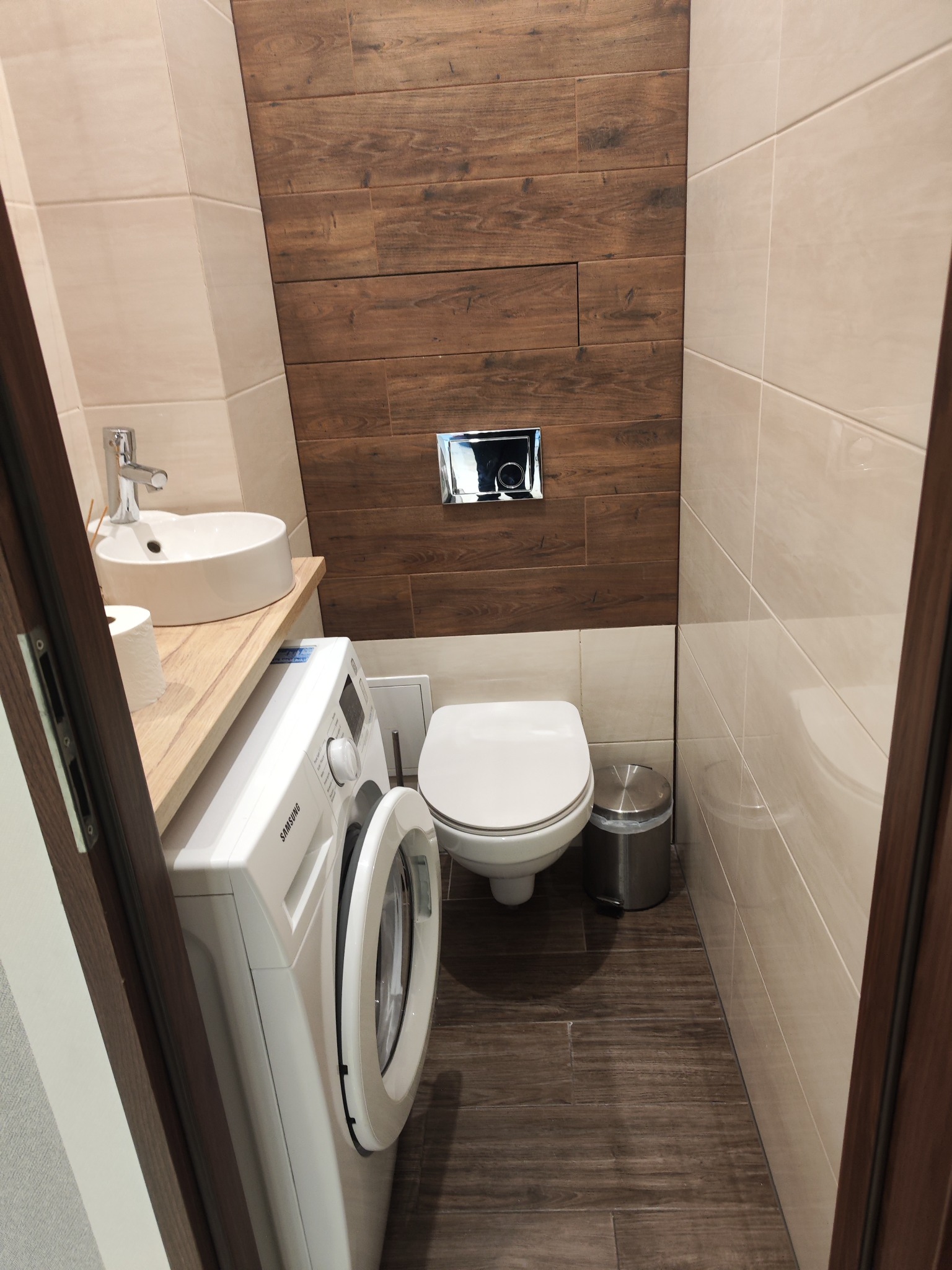 Nowoczesna łazienka z drewnopodobnymi płytkami na ścianie i podłodze, umywalką nablatową na drewnianym blacie, pralką i podwieszaną toaletą.