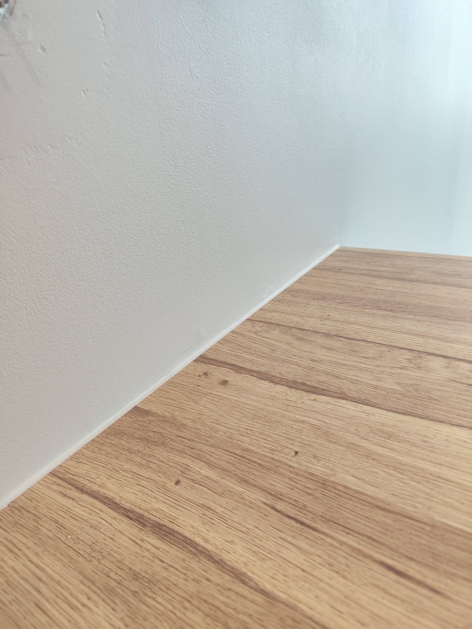 Połączenie gładkiej, pomalowanej na biało ściany i drewnianej podłogi z paneli w jasnym odcieniu dębu, widoczne z bliskiej perspektywy.