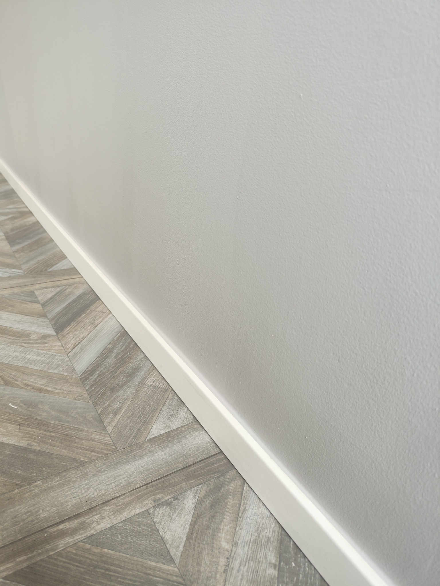 Fragment pokoju po remoncie: jasnoszara ściana pomalowana farbą o matowym wykończeniu, biała listwa przypodłogowa i ułożone panele podłogowe w wzór jodełki francuskiej, imitujące drewno.