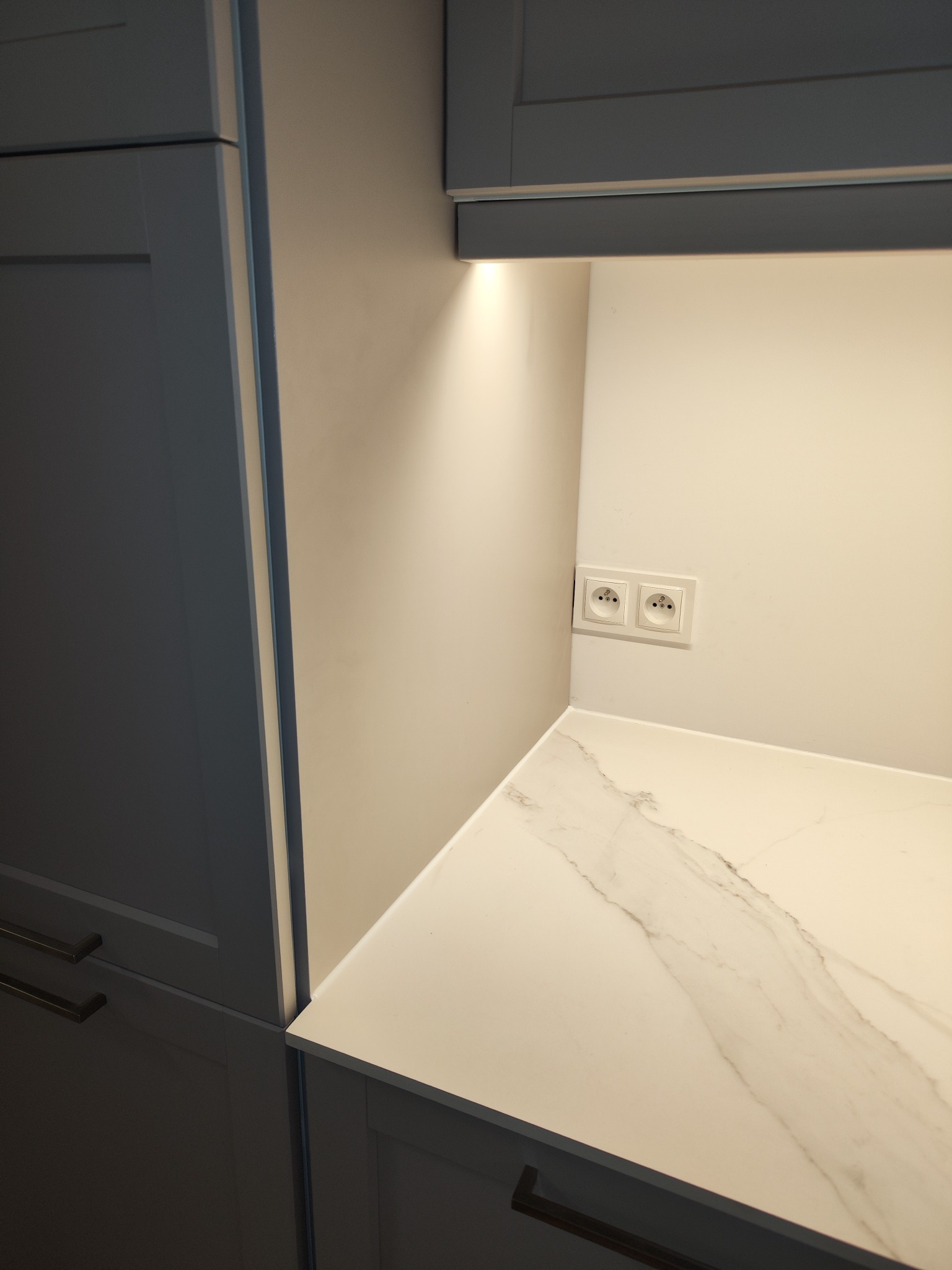 Fragment kuchni po remoncie: szafki w odcieniach szarości, blat z wzorem marmuru, oświetlenie podszafkowe, gniazdka elektryczne w ścianie.
