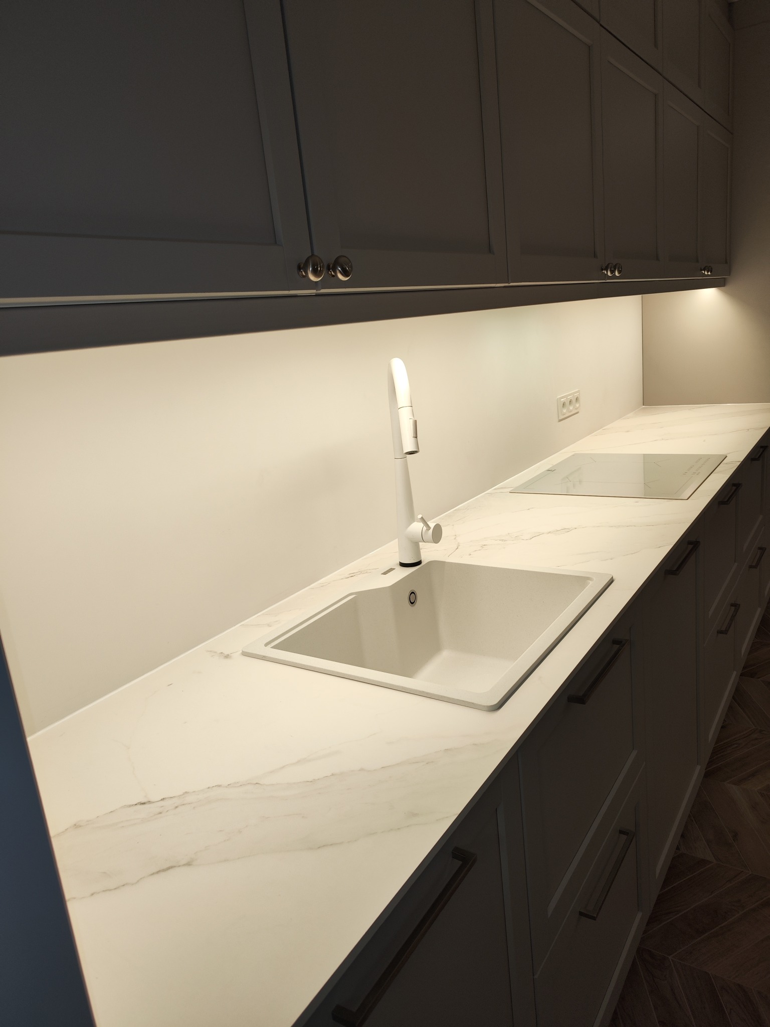 Szara zabudowa kuchenna z białym blatem imitującym marmur, zlewem i baterią w tym samym kolorze, oświetlenie podszafkowe, płyta indukcyjna na blacie.
