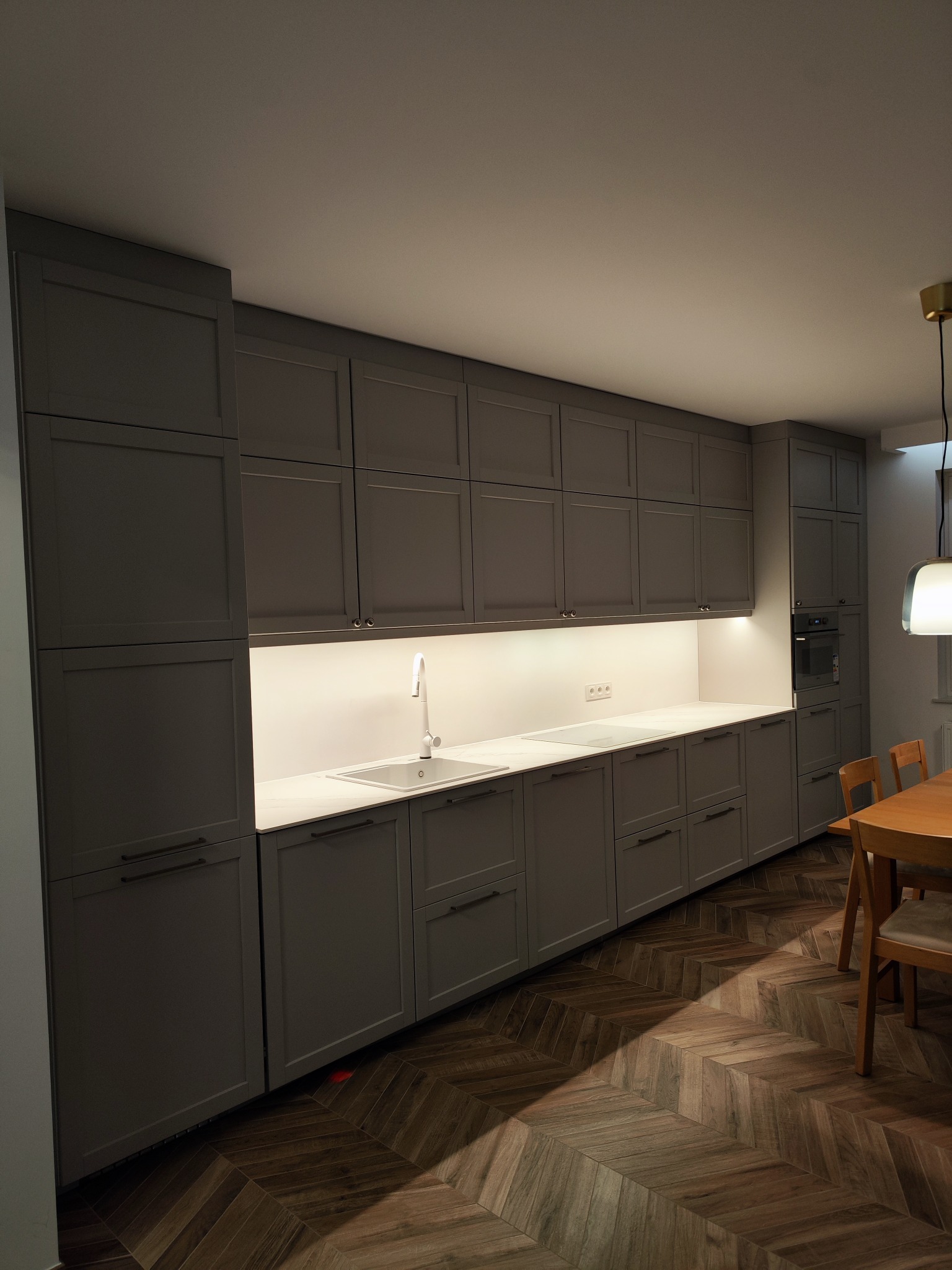 Nowoczesna kuchnia z szarymi, matowymi szafkami, białym blatem, oświetleniem LED pod szafkami wiszącymi i drewnianą podłogą ułożoną we wzór jodełki.