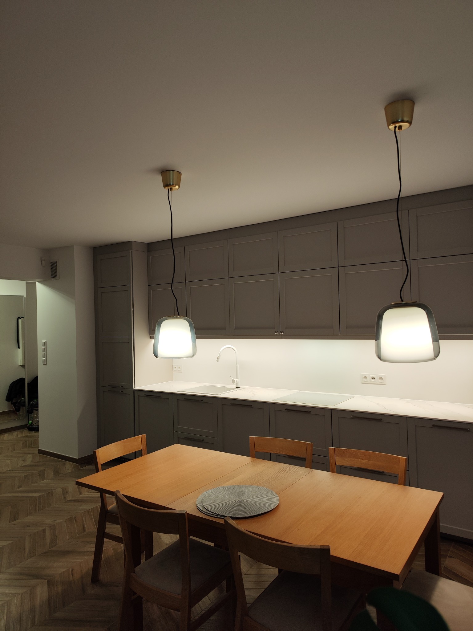 Nowoczesna szara kuchnia z drewnianym stołem i krzesłami, oświetlona dwoma wiszącymi lampami z białymi kloszami i złotymi elementami mocującymi.