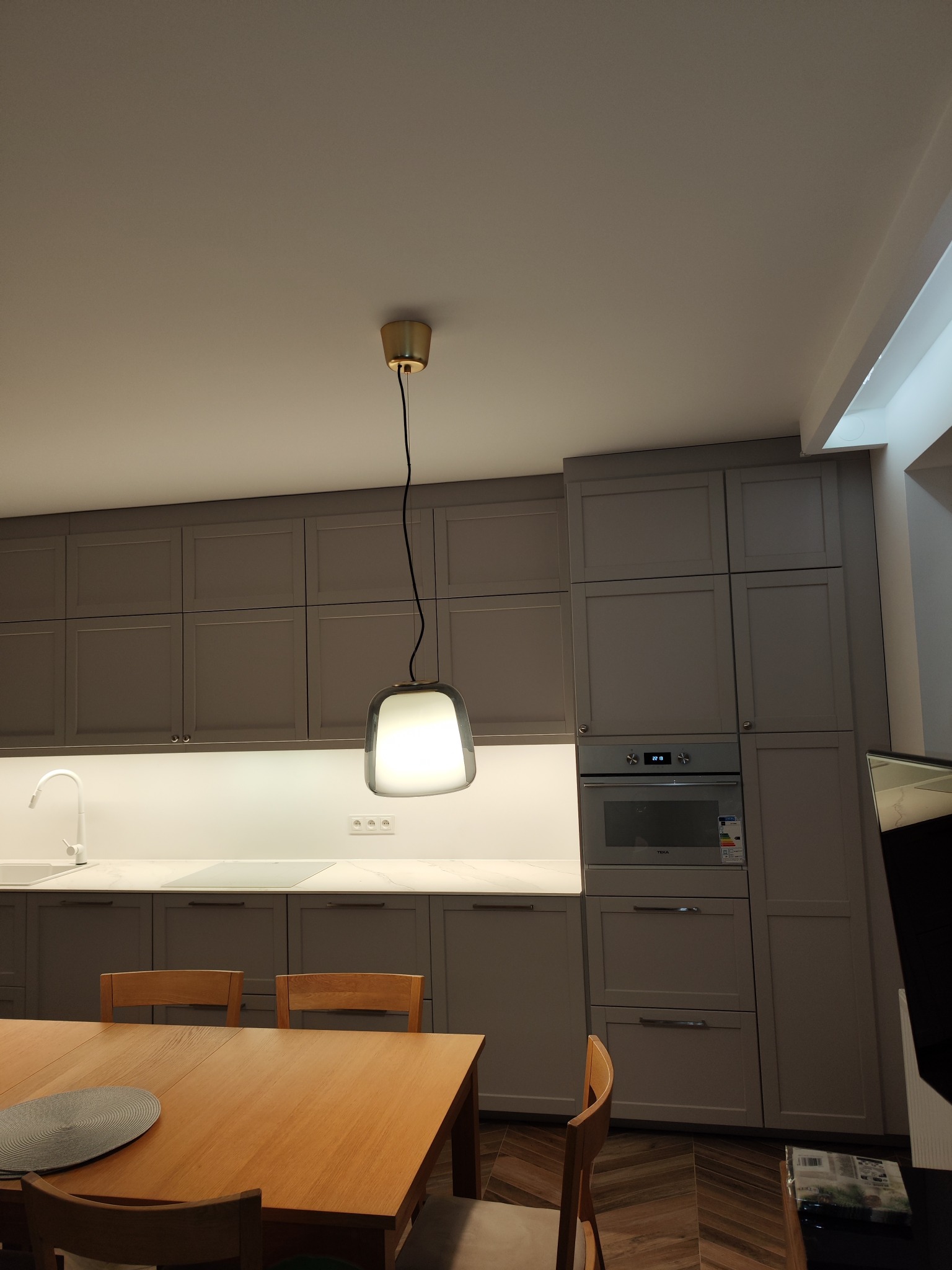 Szare szafki kuchenne z białym blatem, podświetleniem LED i wiszącą lampą z kloszem, widok na kuchenkę i stół z krzesłami.