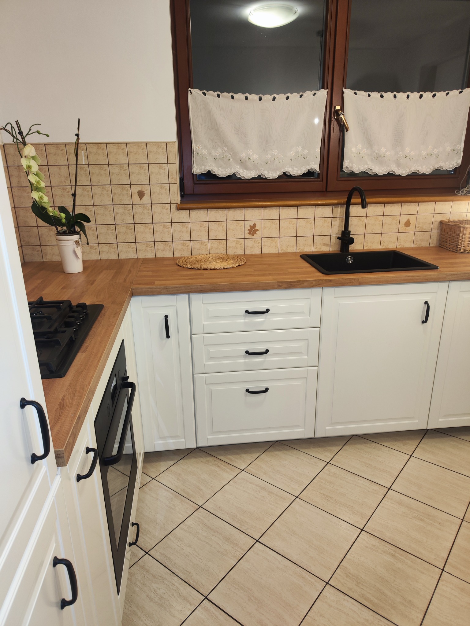 Nowoczesna kuchnia z białymi szafkami, drewnianym blatem, czarną armaturą i płytkami w beżowe wzory na ścianie i podłodze. Na blacie wiklinowa podkładka, a przy oknie białe, haftowane firanki.