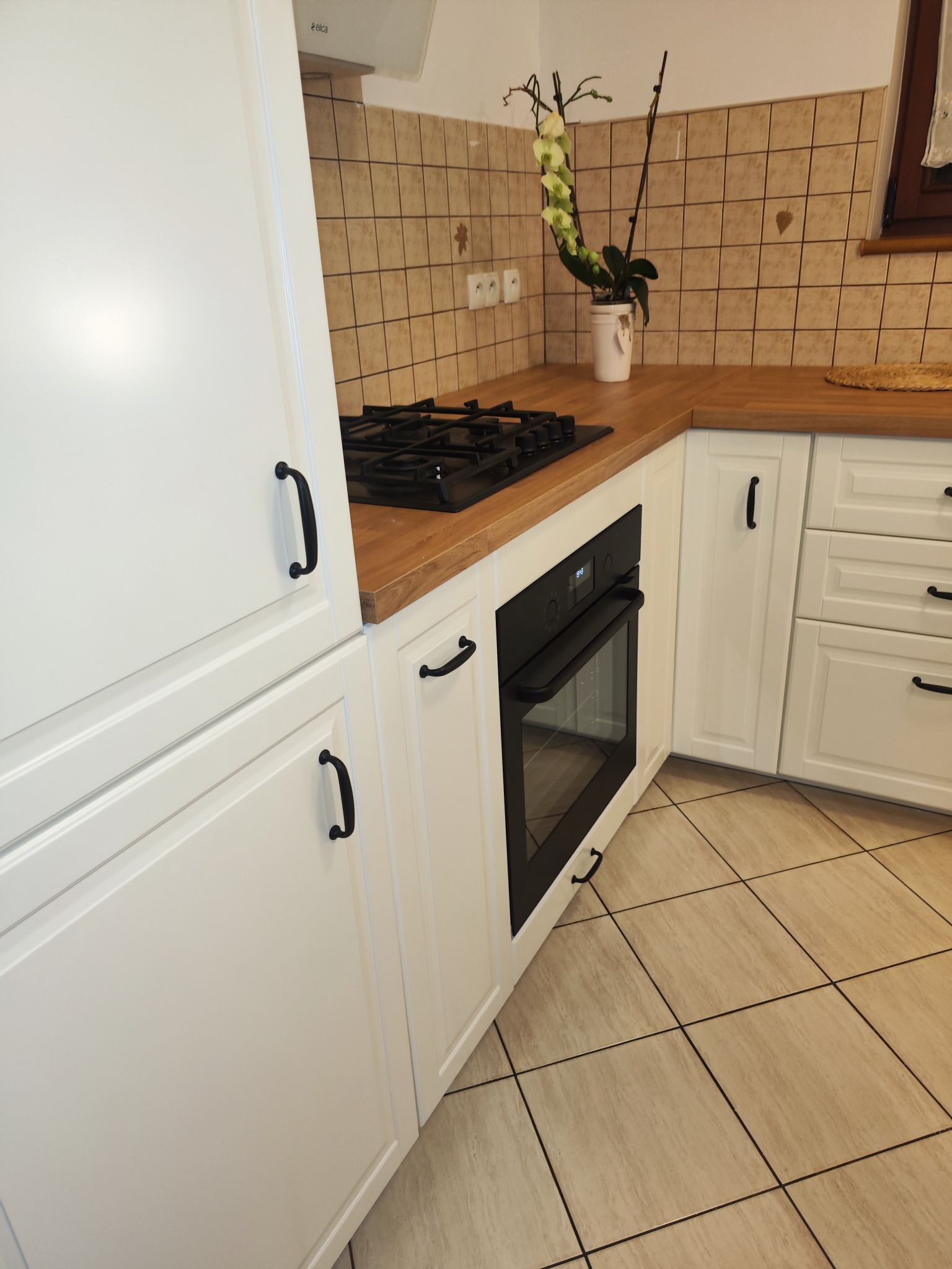Biała zabudowa kuchenna z drewnianym blatem, czarnym piekarnikiem i płytą gazową, zrealizowana w mieszkaniu w Szczecinie. Na blacie stoi doniczka z orchideą.