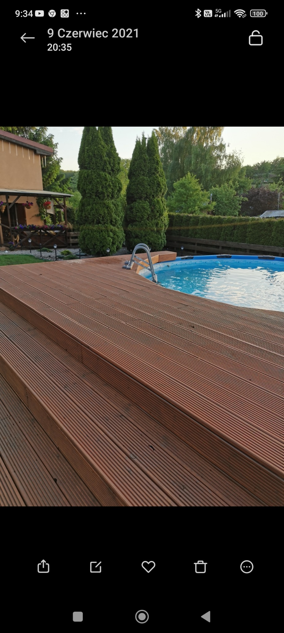 Drewniany taras z ryflowanych desek prowadzący do basenu ogrodowego, widoczne schodki i drabinka do basenu, w tle roślinność ogrodowa i budynek.