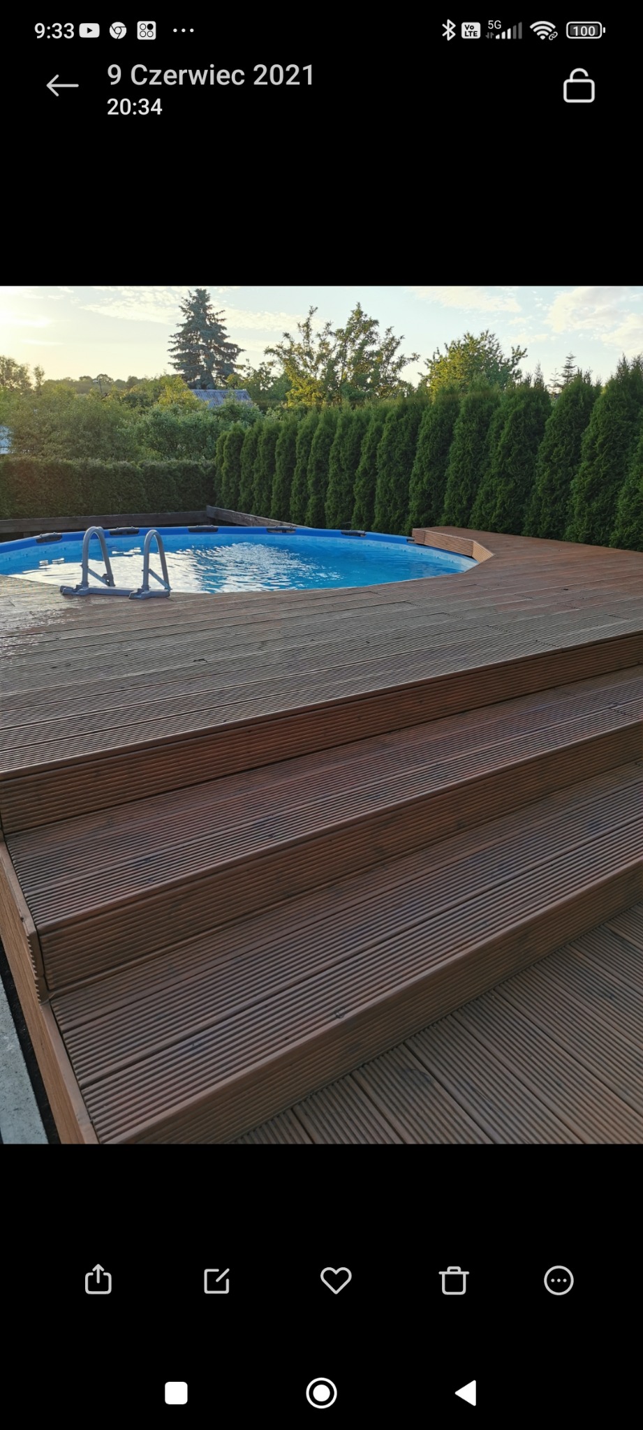 Okrągły basen ogrodowy z metalową drabinką, otoczony drewnianym tarasem z ryflowanych desek i schodami, w tle wysokie tuje.