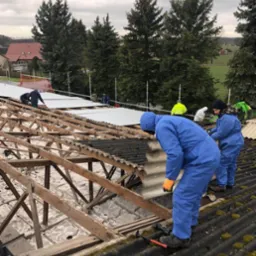 Pracownicy w niebieskich kombinezonach demontują stare, zarośnięte mchem płyty eternitowe z drewnianej konstrukcji dachu, w tle widoczne drzewa i budynki.