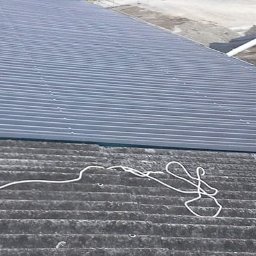 MeisterDach-GmbH-Robert - Fragment dachu z blachy trapezowej w kolorze grafitowym, częściowo położonej na starym, pokrytym mchem dachu z eternitu. Na pierwszym planie biały pojemnik i zwój białego sznurka. W tle widoczny...