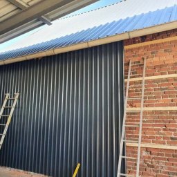 MeisterDach-GmbH-Robert - Montaż blachy trapezowej w kolorze grafitowym na elewacji budynku, widoczna drabina oparta o ścianę z cegły z przygotowanym rusztowaniem pod elewację.