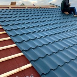 MeisterDach-GmbH-Robert - Montaż ciemnoniebieskiej blachodachówki modułowej na dachu z widoczną konstrukcją więźby i częścią podkładu. Dekarz siedzi na dachu.