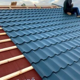 Montaż ciemnoniebieskiej blachodachówki modułowej na dachu z widoczną konstrukcją więźby i częścią podkładu. Dekarz siedzi na dachu.