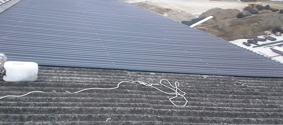 Fragment dachu z blachy trapezowej w kolorze grafitowym, częściowo położonej na starym, pokrytym mchem dachu z eternitu. Na pierwszym planie biały pojemnik i zwój białego sznurka. W tle widoczny...