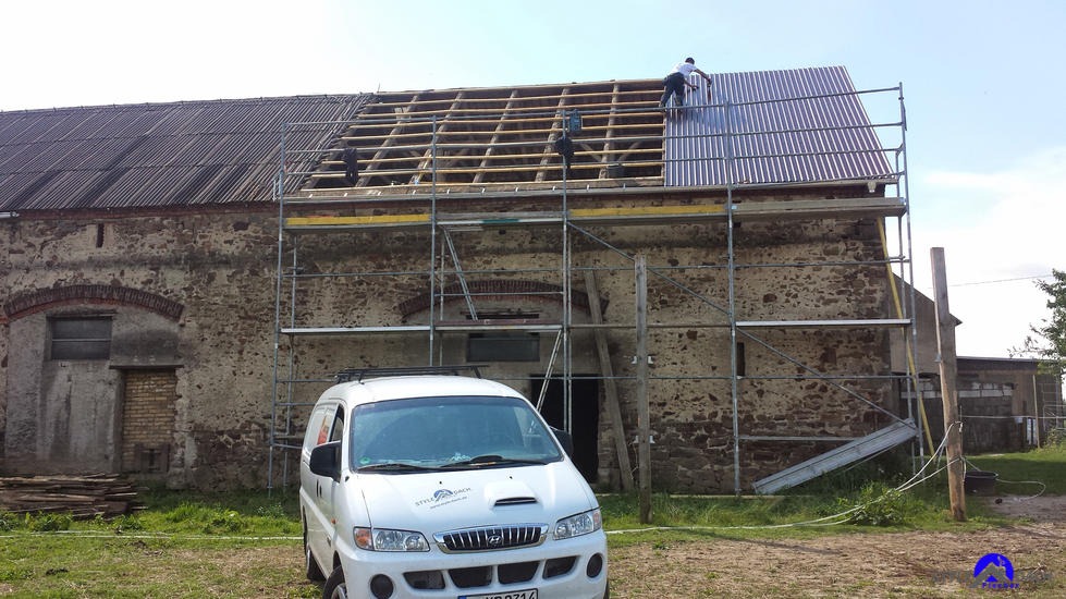 Ekipa dekarzy na rusztowaniu podczas wymiany pokrycia dachu starego budynku gospodarczego, widoczna część drewnianej więźby i fragment nowego, metalowego pokrycia, na pierwszym planie biały...
