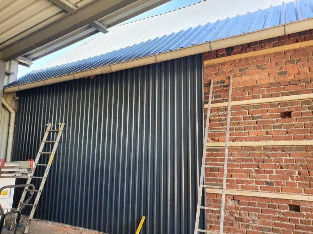 Montaż blachy trapezowej w kolorze grafitowym na elewacji budynku, widoczna drabina oparta o ścianę z cegły z przygotowanym rusztowaniem pod elewację.